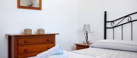 A neatly made bed with a patterned bedspread, a wooden dresser with two spools of thread, a small bedside table with a lamp, and a wall-mounted light fixture.
