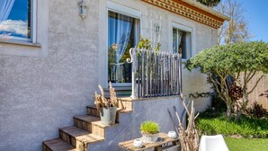 Een huis met een stenen gevel, een houten trap, een metalen hek en een tuin met verschillende planten en een boom.
