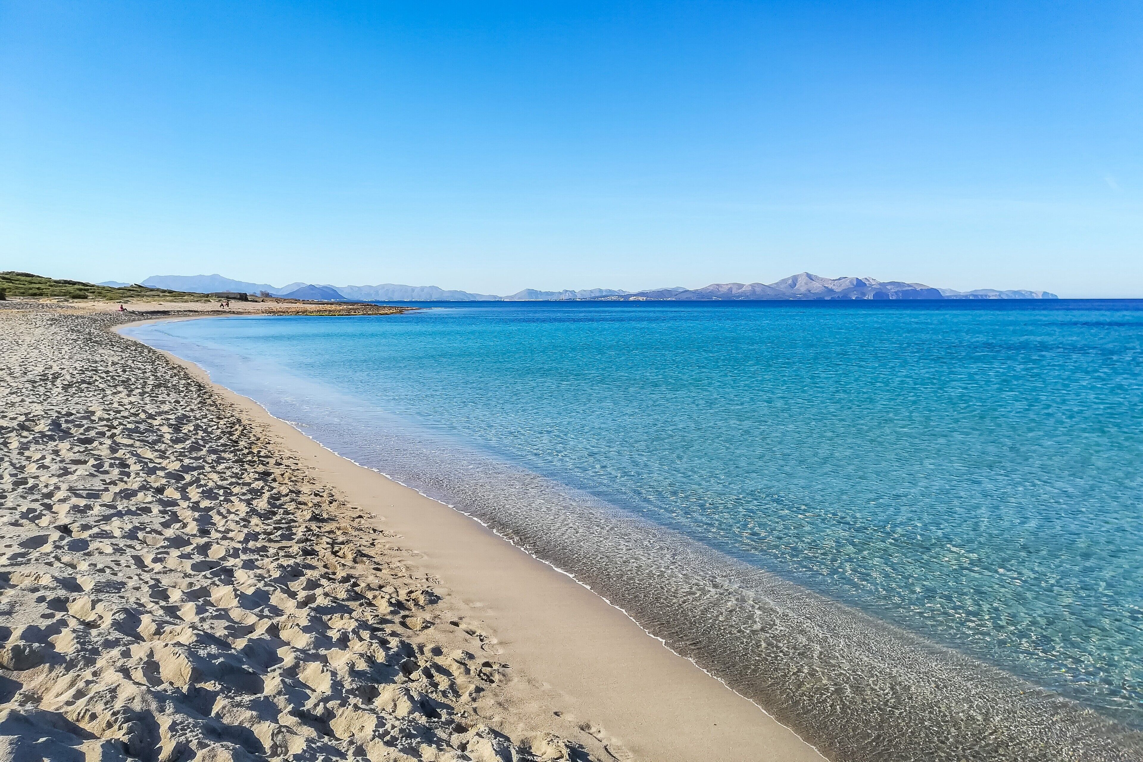 A beach with clear blue water, a sandy shore, and distant mountains.