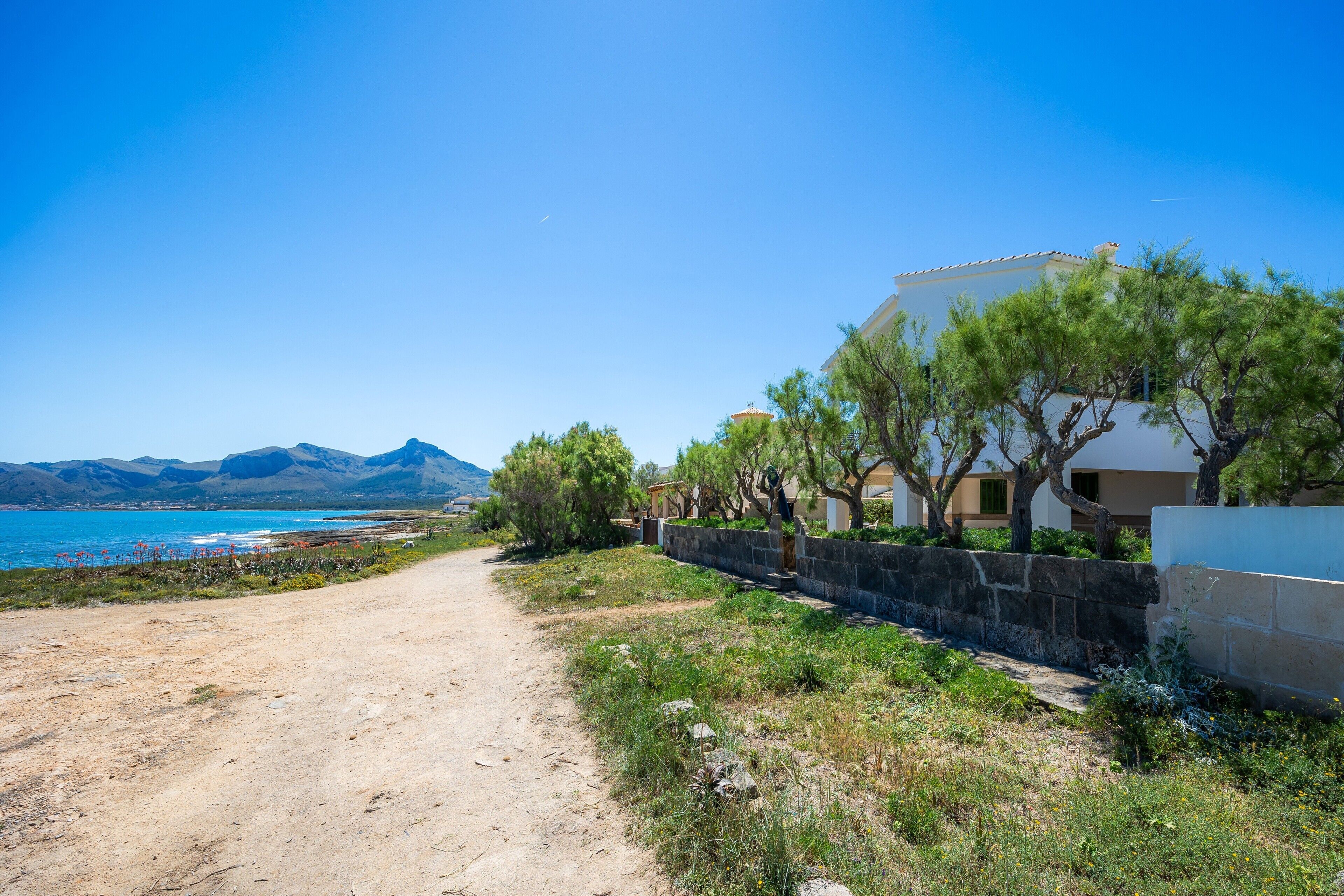 A coastal road leading to a building with a stone wall and trees, overlooking a body of water and mountains.