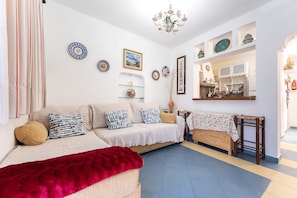 Un salon comprenant un canapé, un plaid rouge, un tabouret en bois et une étagère murale ornée d’assiettes décoratives et d’un tableau encadré.
