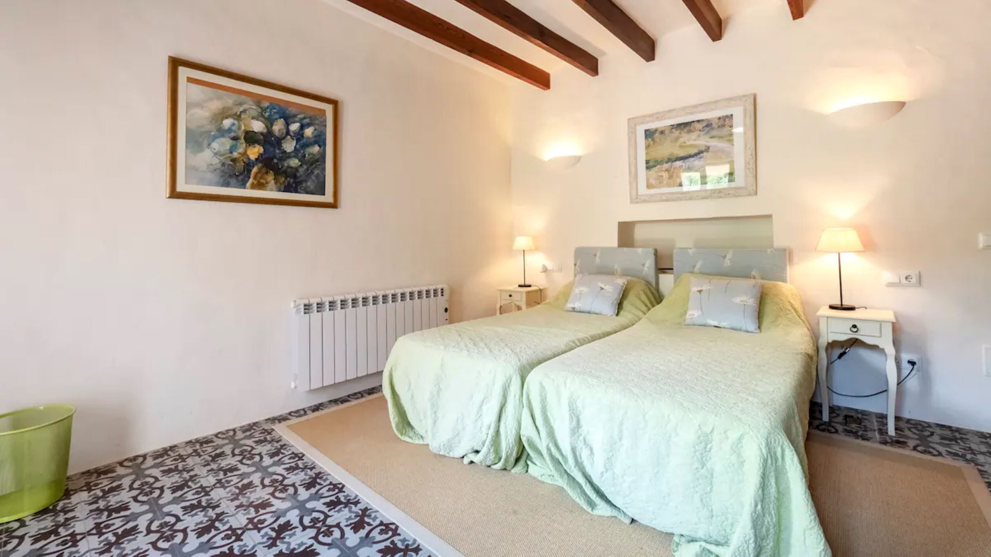 A bedroom with two beds, a radiator, a green trash bin, and artwork on the walls.