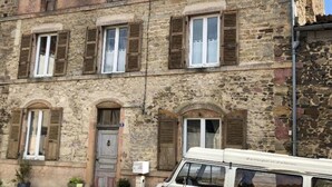 Un bâtiment en pierre de deux étages, doté de nombreuses fenêtres et volets, d’une porte et d’un fourgon rouge garé devant.