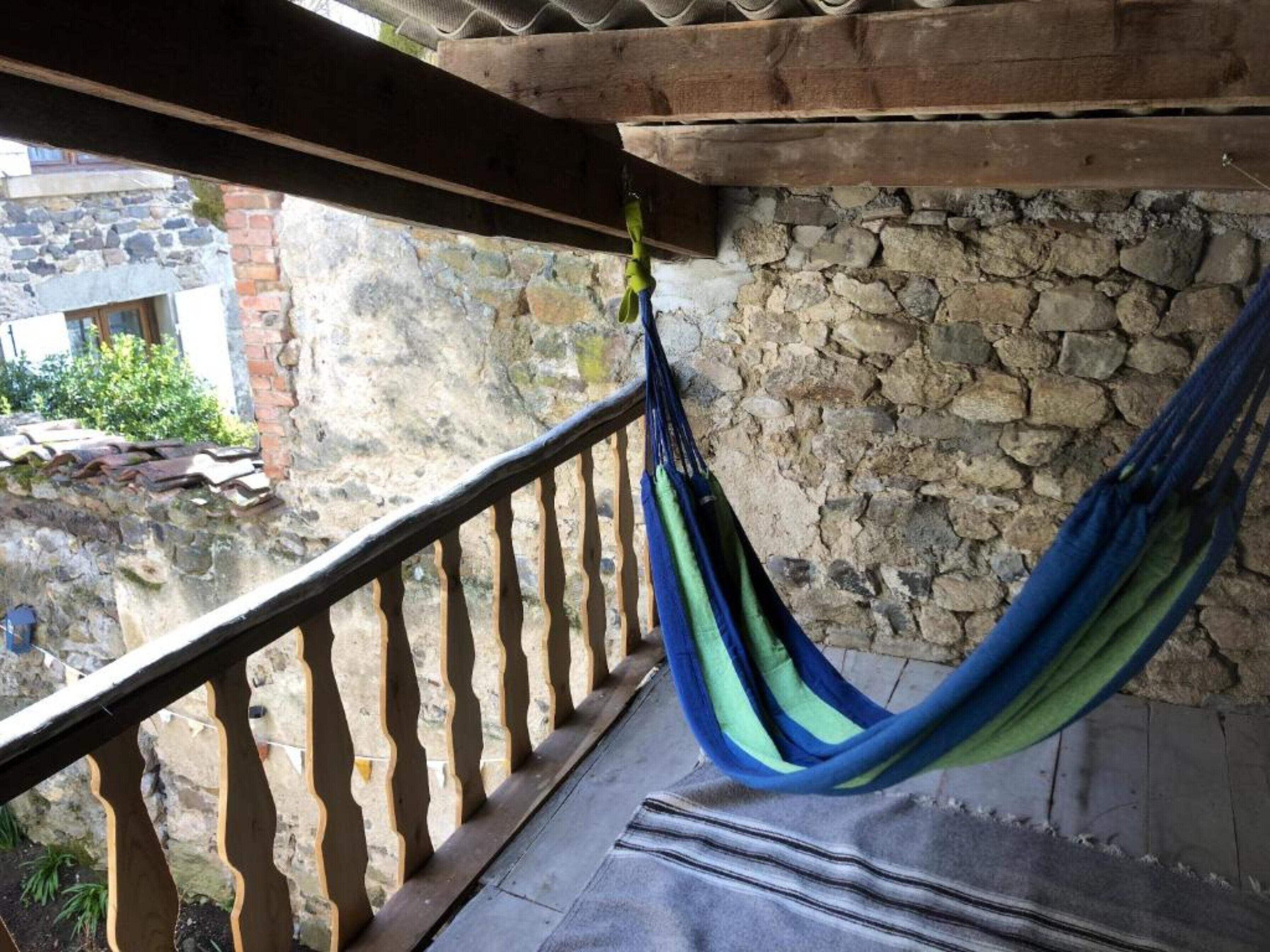 Un hamac est suspendu à la balustrade en bois d’un balcon, avec un mur de pierre en arrière-plan.