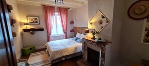 Une chambre à coucher avec un lit, une cheminée, une fenêtre avec des rideaux, un miroir fixé au mur et un chapeau accroché au mur.