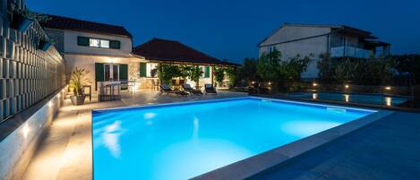 A swimming pool with a tiled edge, surrounded by a patio area with outdoor furniture and a stone wall.