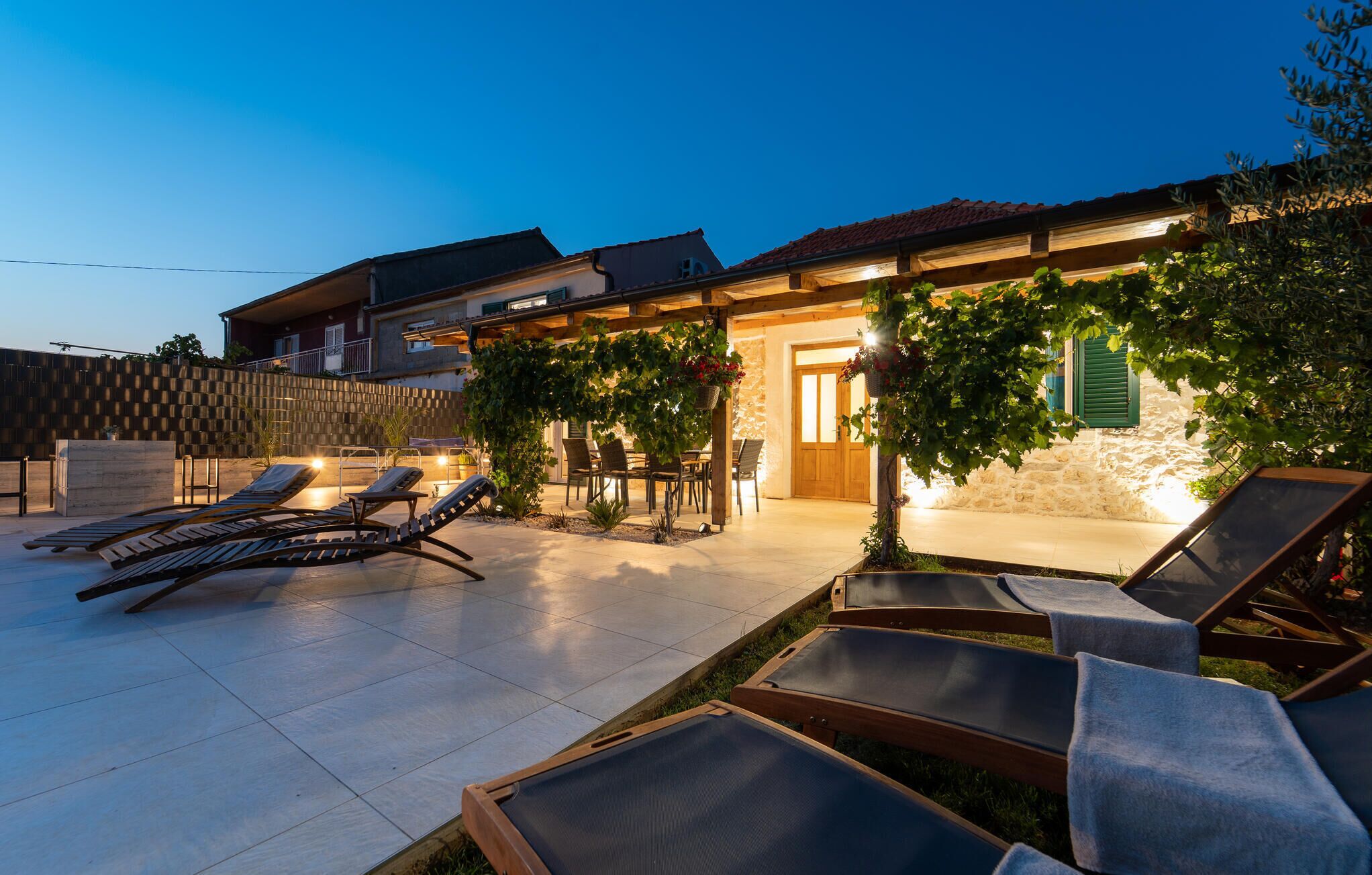 A patio with sun loungers and a dining area, a stone wall, and a wooden pergola with hanging plants.