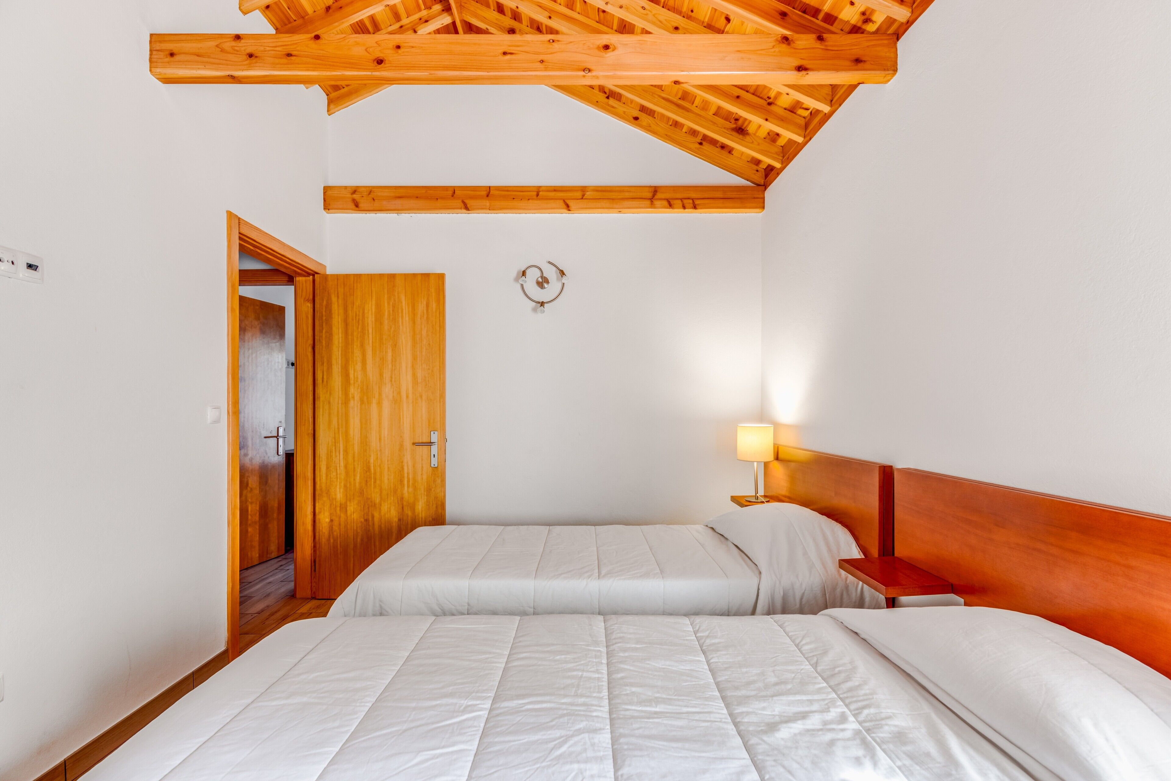 A bedroom with two beds, wooden ceiling beams, a nightstand with a lamp, and a door leading to another room.
