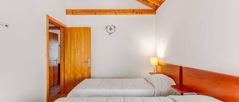 A bedroom with two beds, wooden ceiling beams, a nightstand with a lamp, and a door leading to another room.