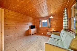 A wooden cabin interior with a sofa, a television on a wooden cabinet, a window with curtains, and a radiator.