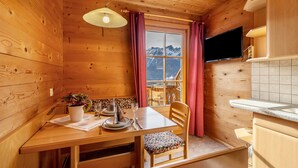 Interior de uma cabana de madeira com uma mesa de jantar, cadeiras, um vaso de planta, uma TV na parede e uma janela com cortinas.