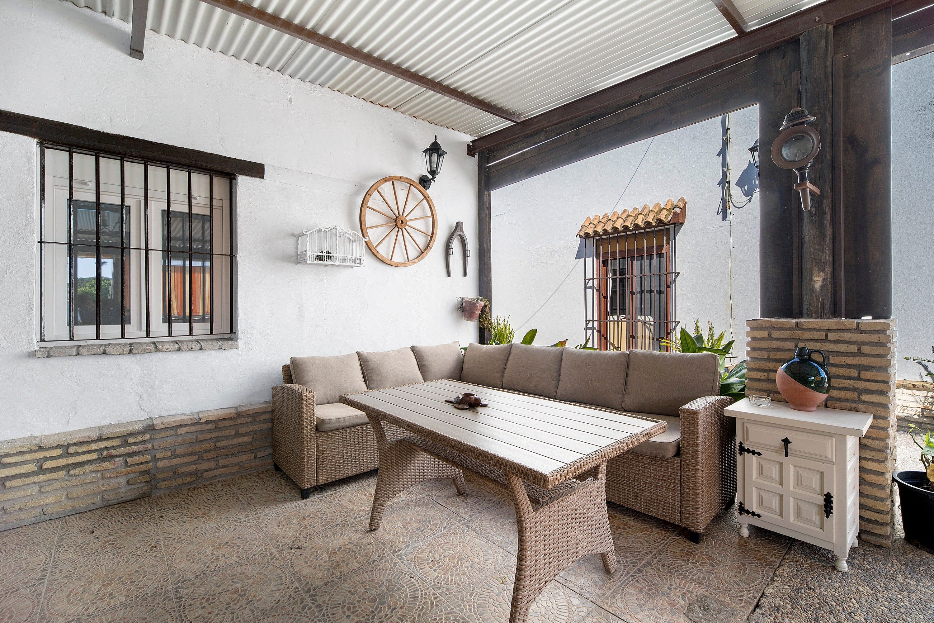 Un patio techado con un sofá de mimbre, una mesa de madera y un armario blanco.