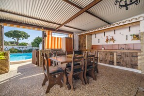An outdoor patio with a pool, a dining area with wooden furniture, and a kitchen area with a brick wall and hanging utensils.
