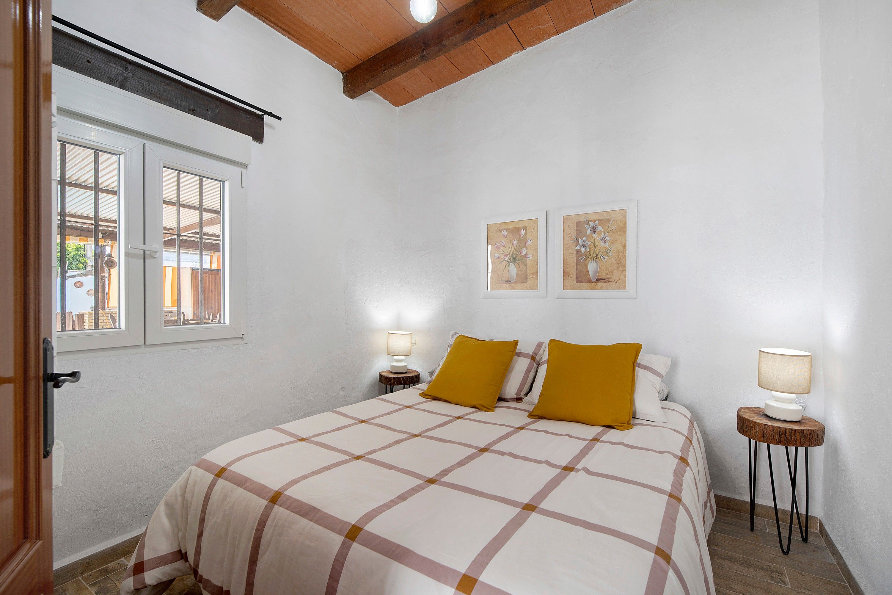 A bedroom with a bed, two bedside lamps, a window with a view, and two wall-mounted pictures.