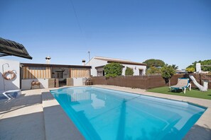 A swimming pool with a clear blue water surface, surrounded by a white building with a tiled roof and a brown wall.