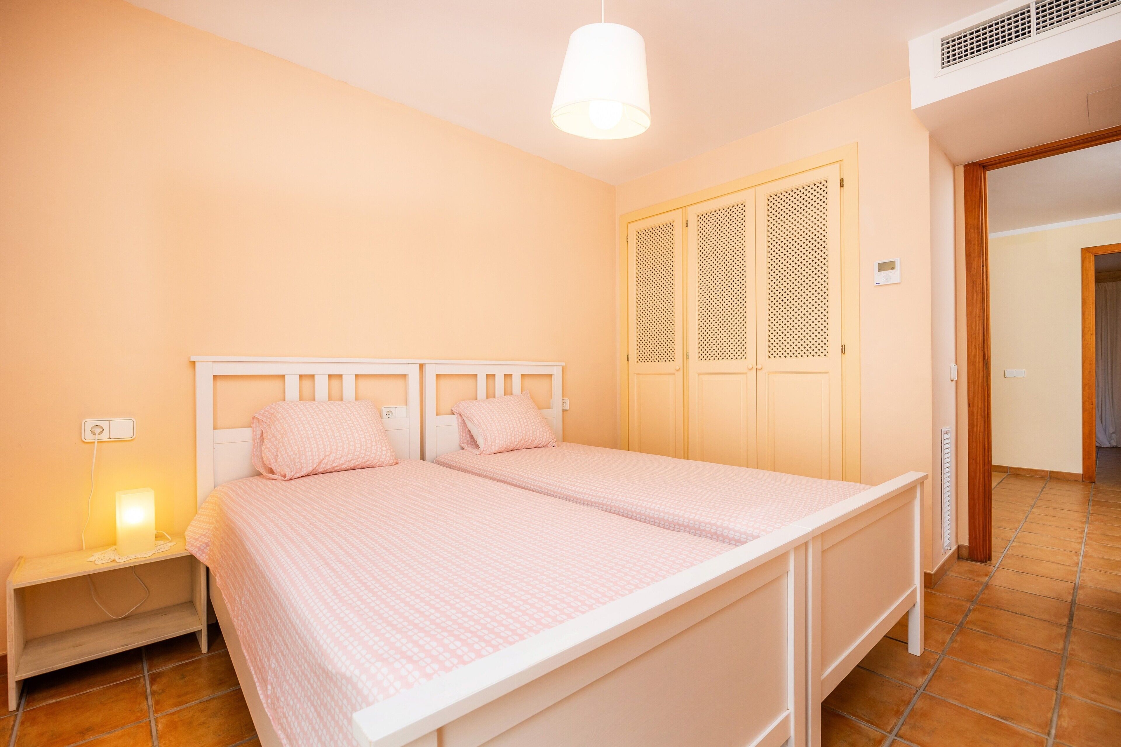 Un dormitorio con cama doble, una mesita de noche con lámpara, un armario con puerta persiana y una rejilla en el techo.
