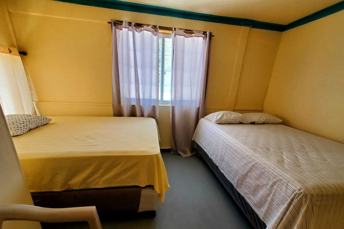A small, single-room hotel room with two beds, a chair, and a window with curtains.