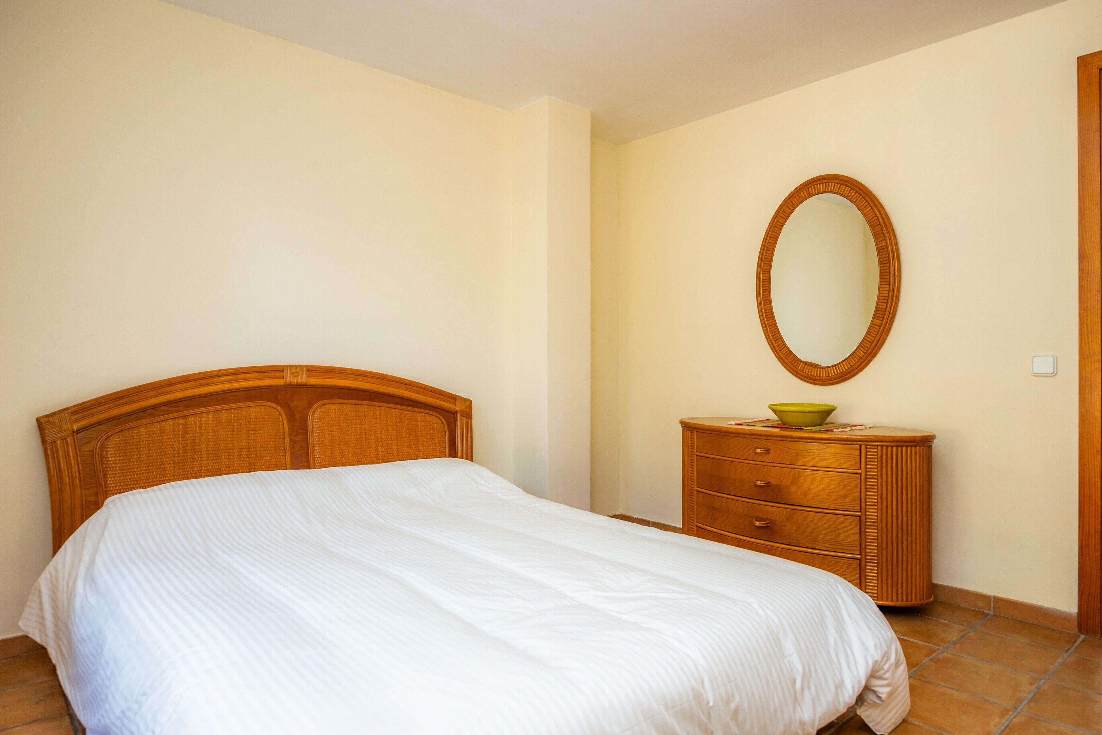 A bedroom with a wooden headboard, a bed with white bedding, a wooden dresser with a mirror, and a green bowl on top.