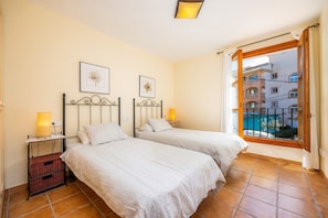 A bedroom with two beds, a window with a view of buildings, and a small table with a lamp.