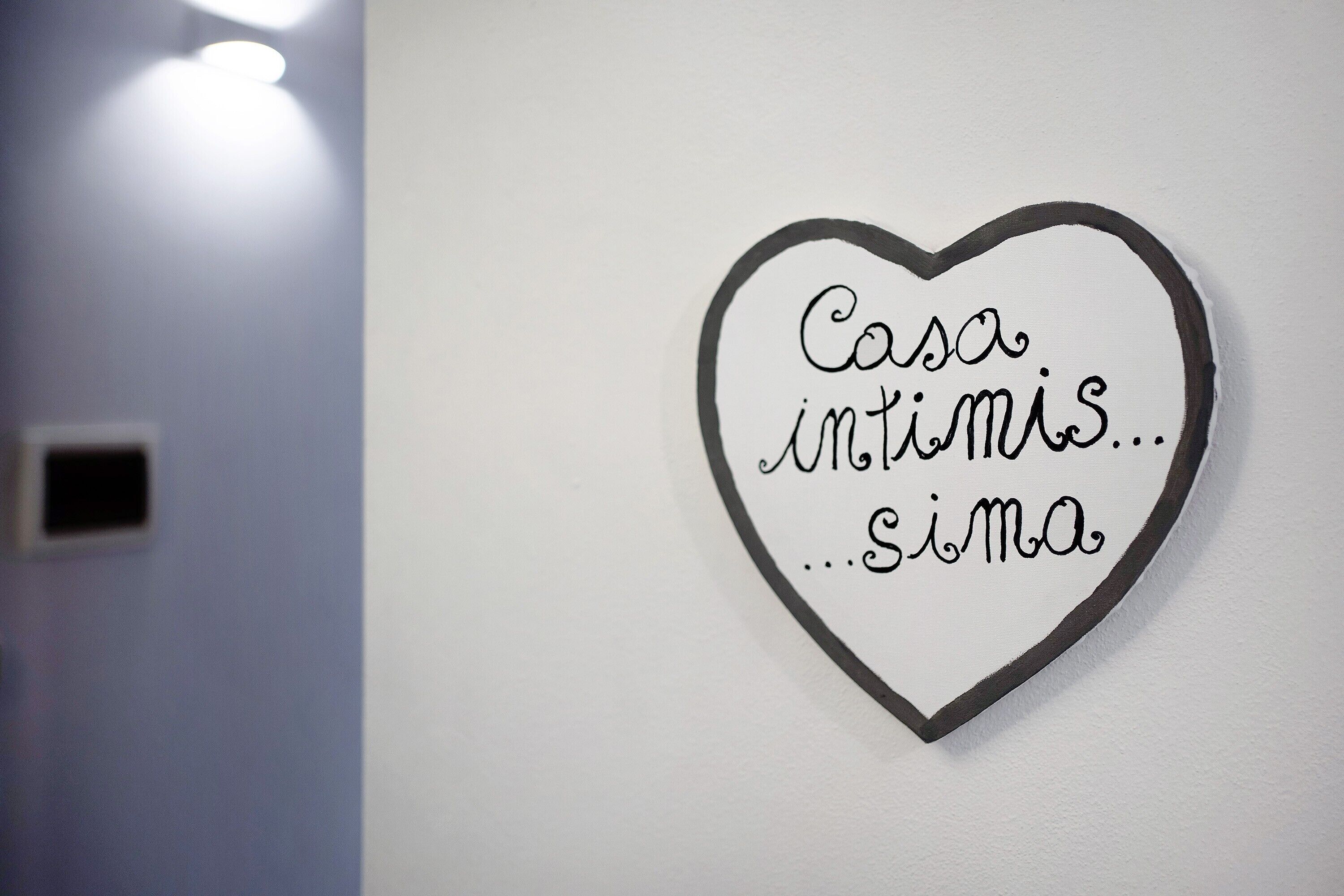 Un cartello a forma di cuore con il testo 'Casa intimis...simma' su una parete bianca.