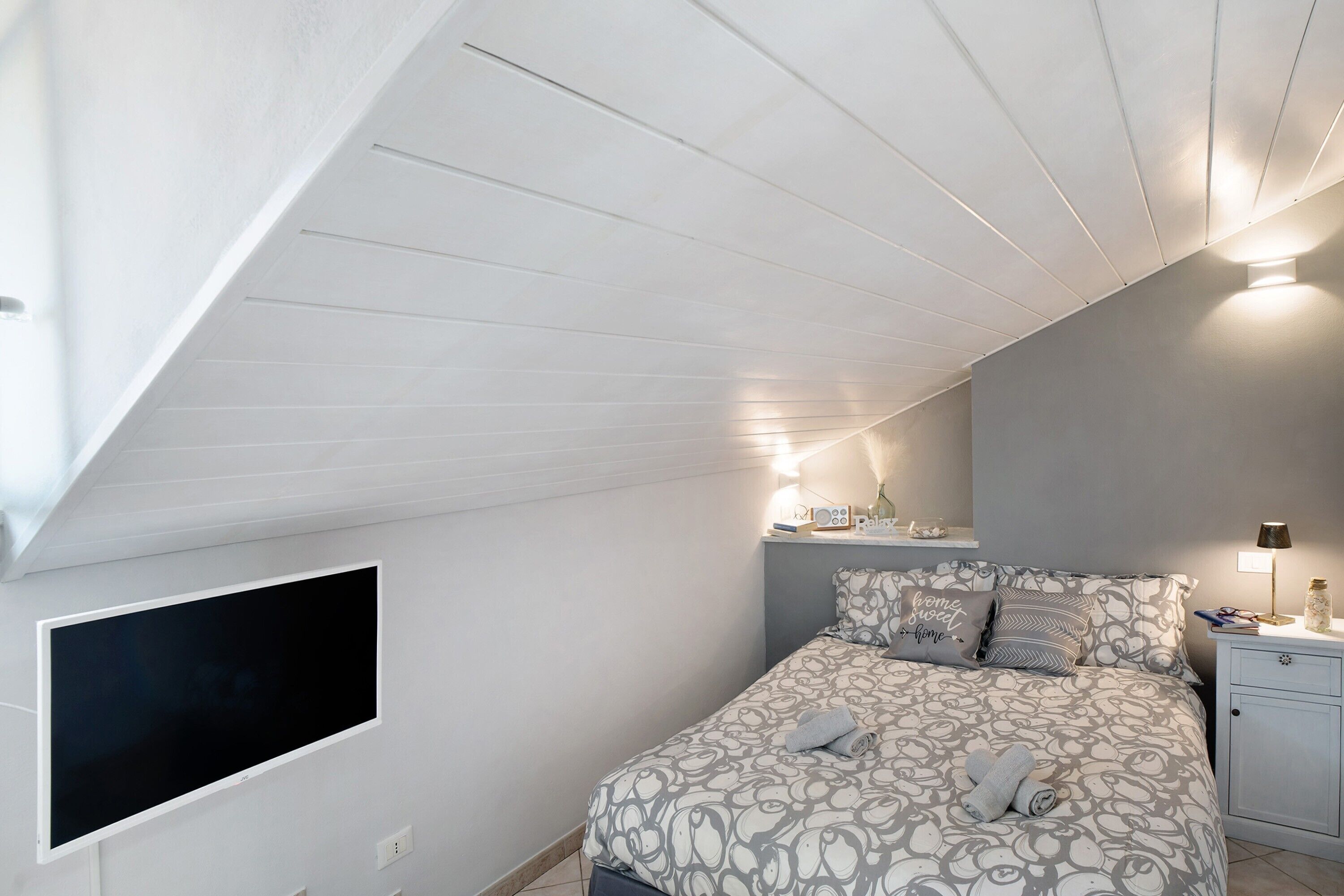 Une chambre avec un plafond en pente, un lit avec une literie à motifs, une table de chevet avec une lampe et un téléviseur à écran plat fixé au mur.