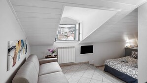 A bedroom with a sloped ceiling, a bed, a sofa, a radiator, a TV, and a window with a view of buildings.