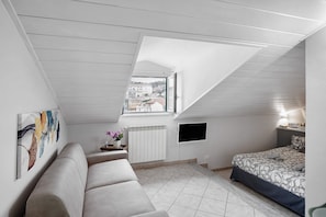 A bedroom with a sloped ceiling, a bed, a sofa, a radiator, a TV, and a window with a view of buildings. - Holiday Apartment 'Casa Intimissima' with Air Conditioning (Imperia)