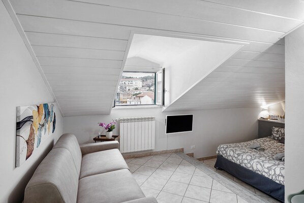 A bedroom with a sloped ceiling, a bed, a sofa, a radiator, a TV, and a window with a view of buildings. - Holiday Apartment 'Casa Intimissima' with Air Conditioning (Imperia)