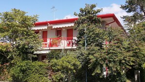 Ein zweistöckiges Gebäude mit rotem Dach und Balkon, umgeben von üppiger Bepflanzung.