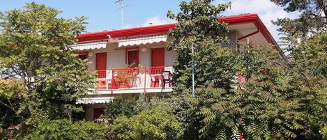 A two-story building with a red roof and balcony, surrounded by dense greenery.