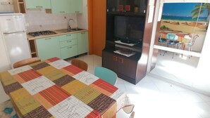 A kitchen with a refrigerator, stove, sink, and cabinets. There's a dining table with a colorful tablecloth and chairs. A TV is mounted on the wall, and a window with a beach view is visible.