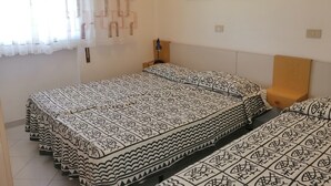 A room with two beds, a painting of boats, a lamp, and a window with patterned curtains.