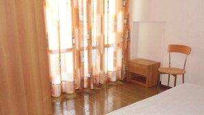 Un dormitori amb un llit, una cadira, una taula petita i cortines.