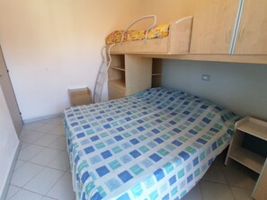 A small, single-bed room with a bunk bed above, a wooden wardrobe, a small desk, and a tiled floor.
