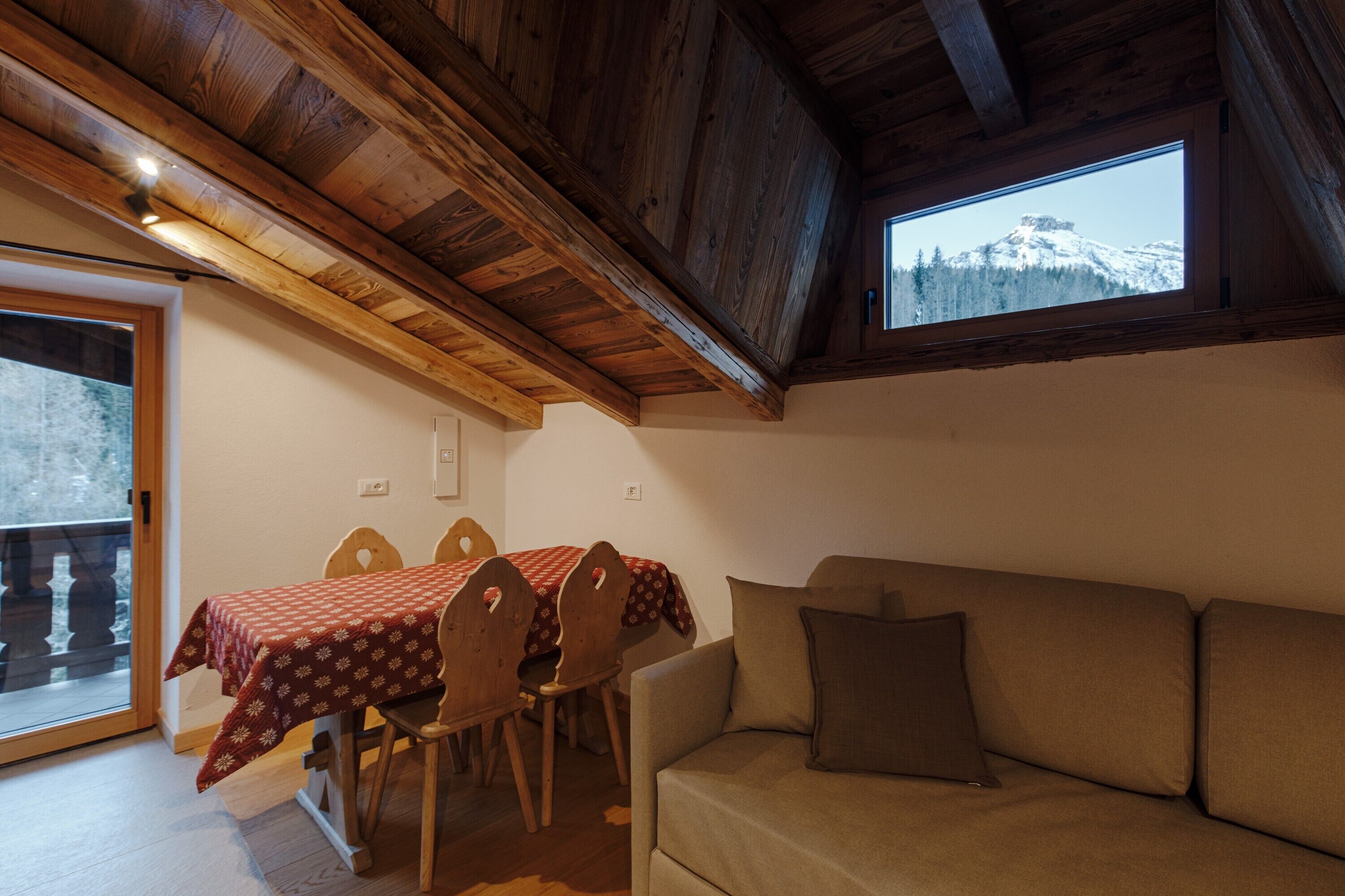 Un comedor con una mesa y sillas, un sofá, vigas de techo de madera y una ventana con vistas a la montaña.