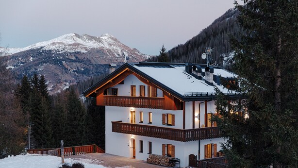 A two-story chalet with a balcony, a wooden deck, and a mountain backdrop.