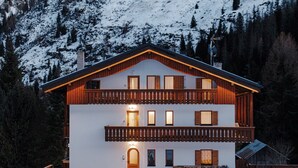A two-story chalet with a balcony, set against a snowy mountain backdrop.