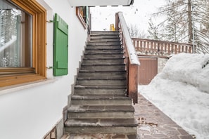 Un escalier menant à une porte en bois, une fenêtre avec des volets verts et une zone recouverte de neige.