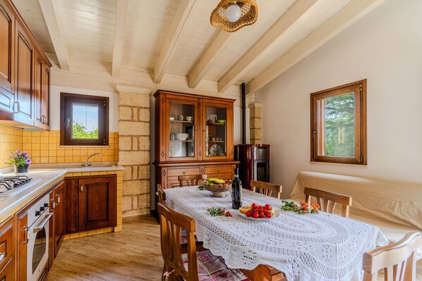 Une cuisine avec des meubles en bois, une table à manger recouverte d’une nappe en dentelle et une vue sur l’extérieur par une fenêtre.