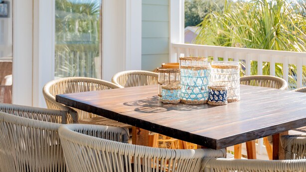 Une terrasse couverte avec une table en bois et des chaises en osier, agrémentée d’un luminaire suspendu doré et de pots décoratifs.