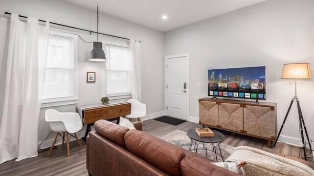 A modern living room with a brown leather sofa, a small round table, a white chair, a wooden cabinet, and a TV mounted on the wall. - Gorgeous Getaway -1BR- Columbus! (Columbus)