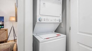 A compact, modern laundry room with a front-loading washer and dryer, a small table with a lamp, and a television on the wall. - Gorgeous Getaway -1BR- Columbus! (Columbus)