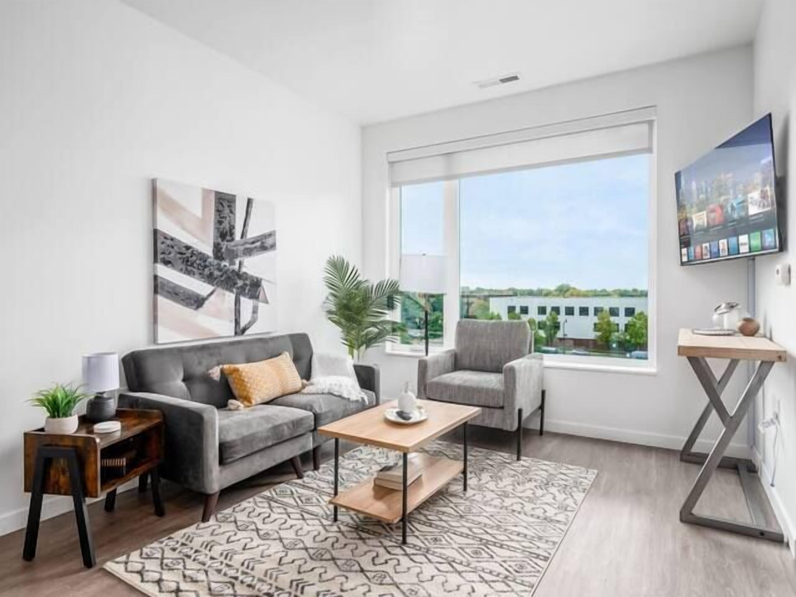 Ein modernes Wohnzimmer mit einem grauen Sofa, einem Holztisch und einem grauen Sessel. Ein großes Fenster bietet Blick auf Gebäude und Grünflächen.