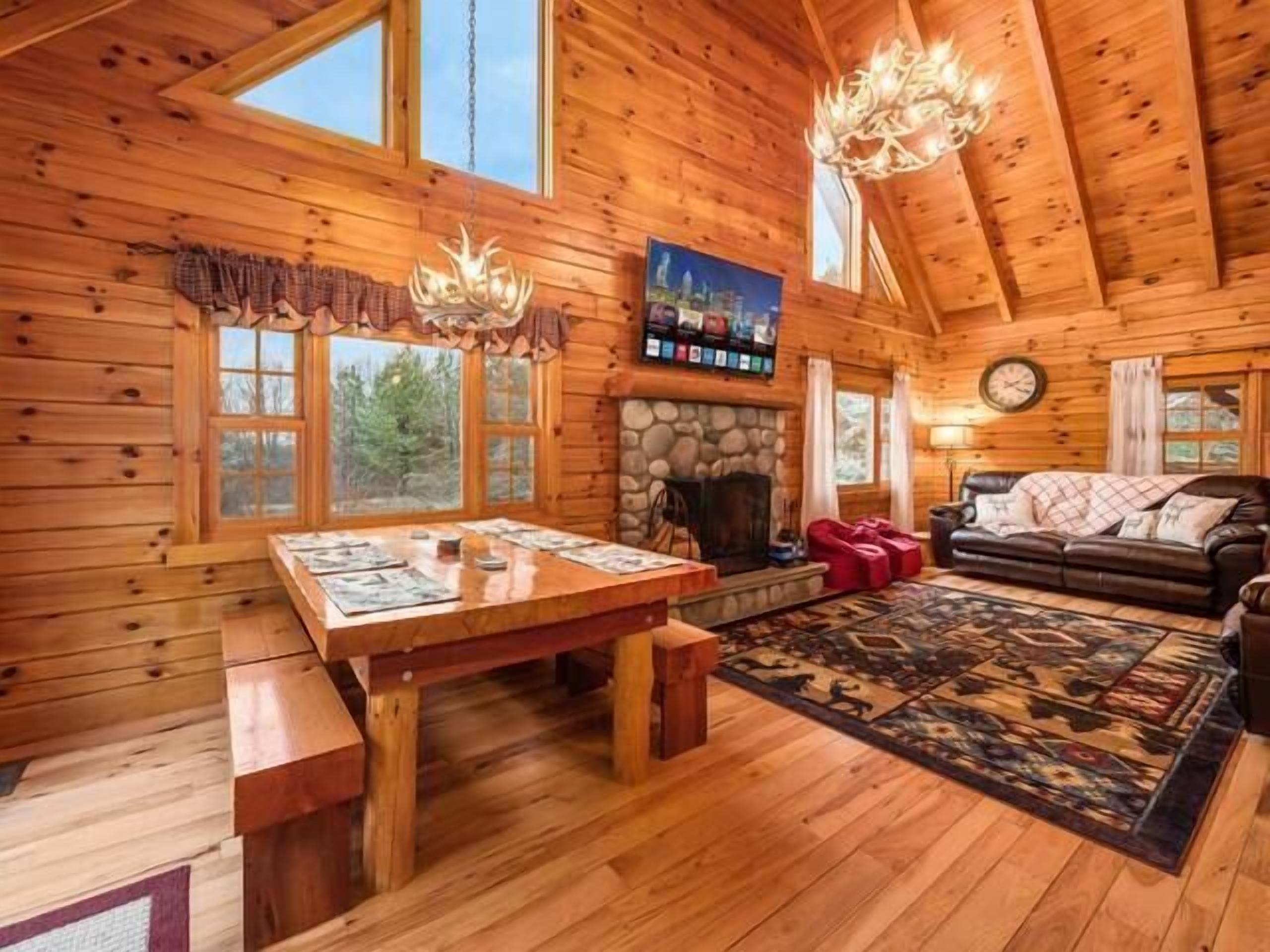 A log cabin interior with a stone fireplace, wooden dining table, and a cozy living area with a sofa and rug.
