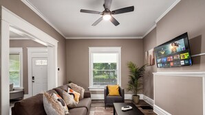A living room with a brown sofa, a gray armchair, a coffee table, and a TV mounted on the wall displaying various channels. - Perfect Stay - 2 Bedrooms - Columbus! (Columbus)