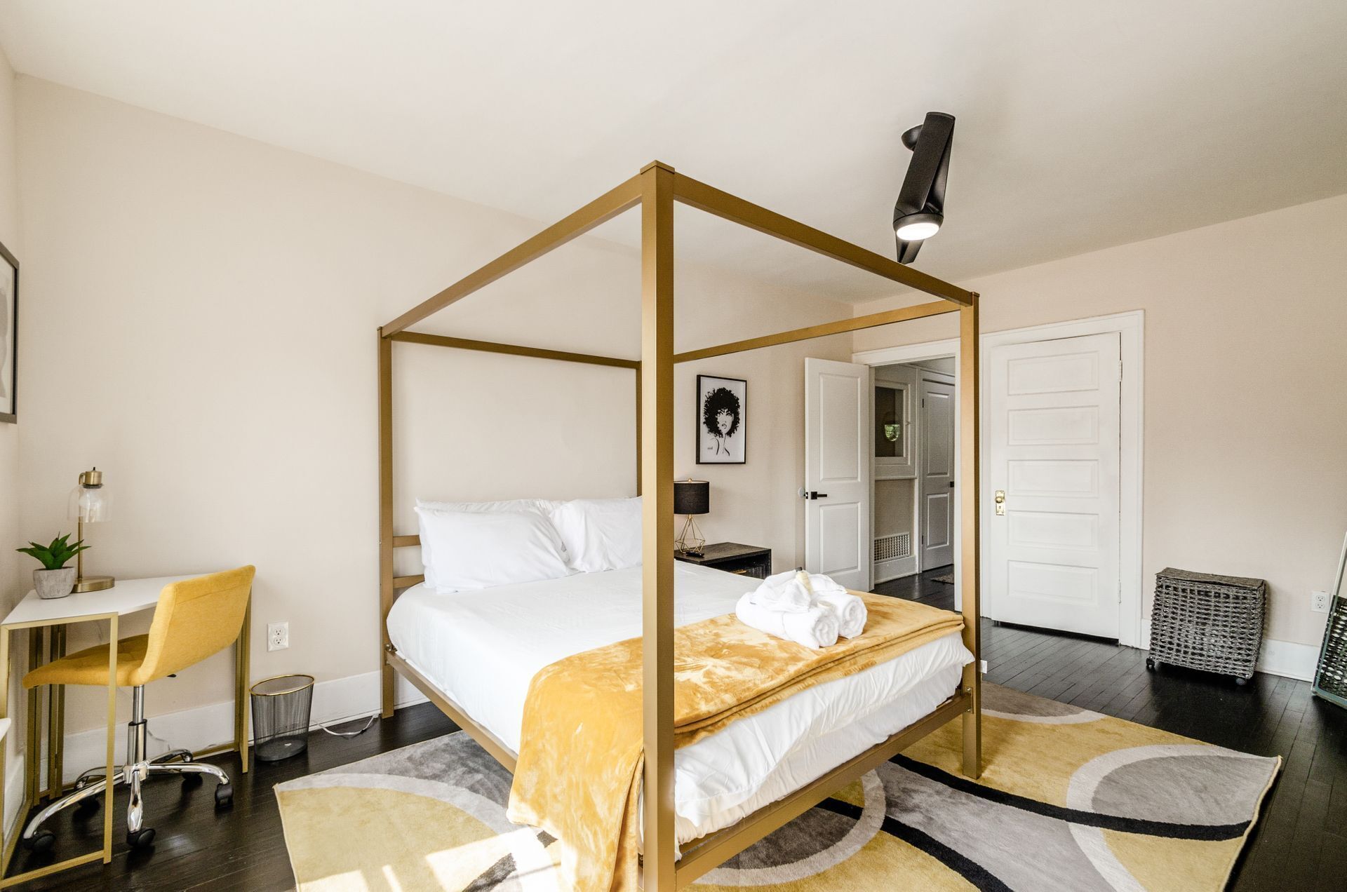 Un lit à baldaquin avec une couvre-lit blanche et un plaid jaune, un bureau avec une chaise, une plante en pot et une lampe.