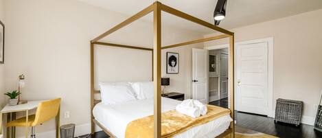 Un lit à baldaquin avec une couvre-lit blanche et un plaid jaune, un bureau avec une chaise, une plante en pot et une lampe.
