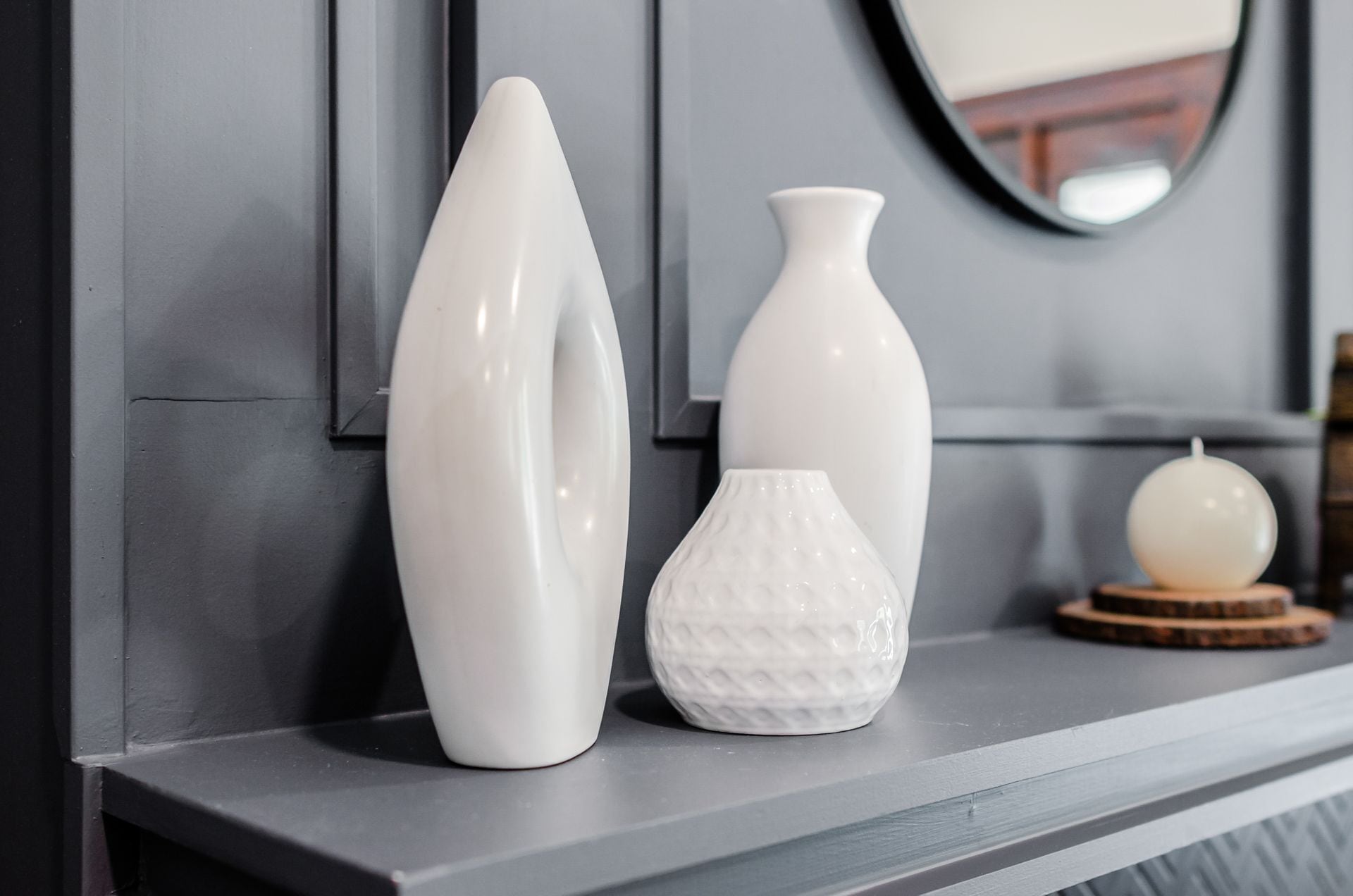 Trois vases blancs sont posés sur une étagère grise, adossée à un mur gris, et un miroir rond est accroché au-dessus.
