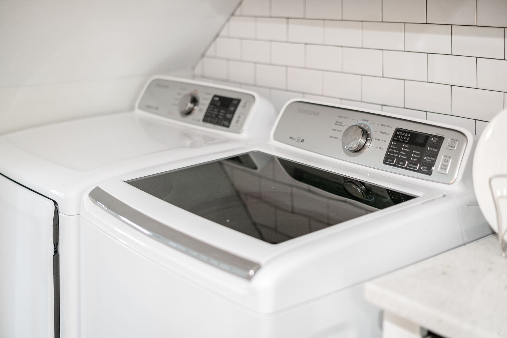 A front-loading washing machine with a control panel, set in a kitchen with white tiled walls.