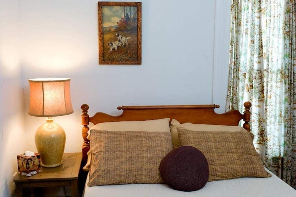 A bedroom with a wooden bed, two pillows, a round pillow, a lamp, a painting, and floral curtains.
